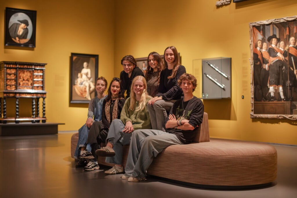 Groep mensen zittend op een houten bank in een museumzaal met gele wanden en kunstwerken.