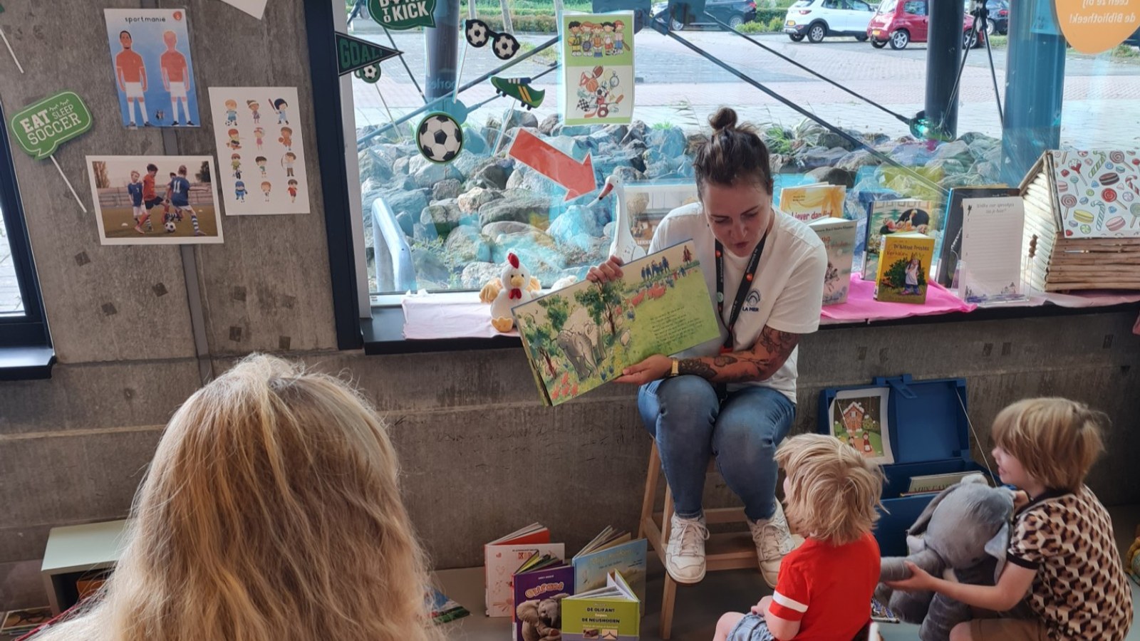 Bibliothecaris leest prentenboek voor aan groep peuteers zittend op vloer van bibliotheek met speelgoed en posters op muur