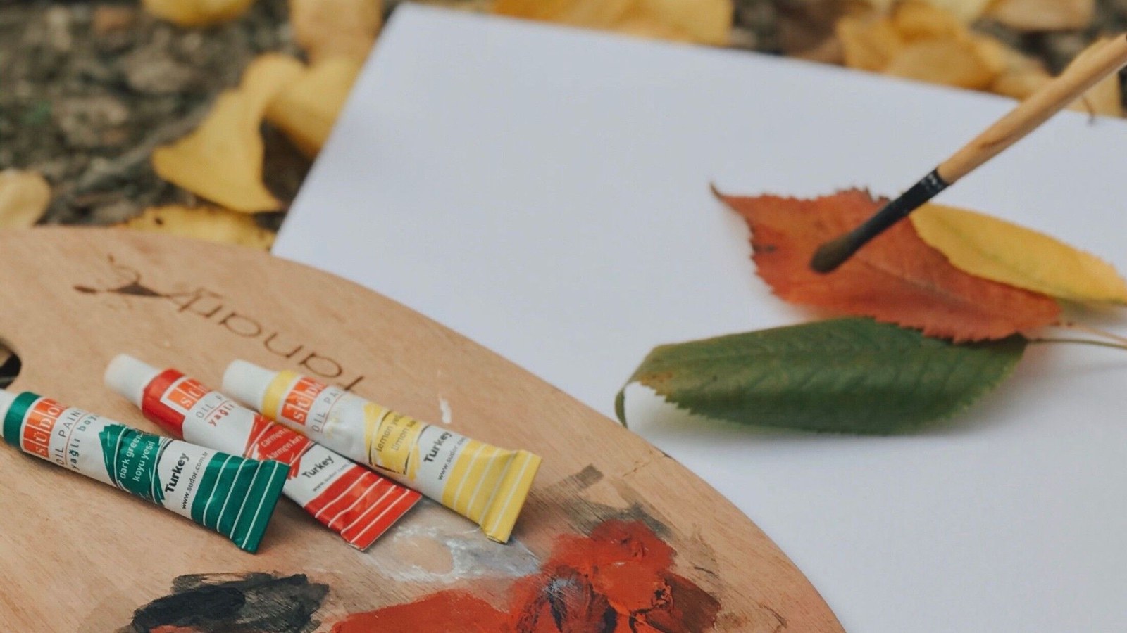 Houten palet met verftubes en penseel, blanco papier en gekleurde bladeren op werkblad
