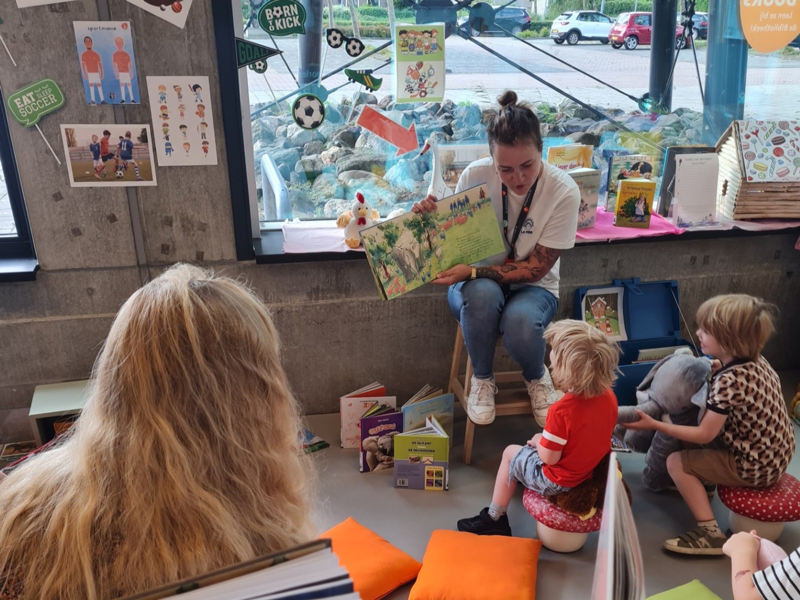 Bibliothecaris leest prentenboek voor aan groep peuteers zittend op vloer van bibliotheek met speelgoed en posters op muur