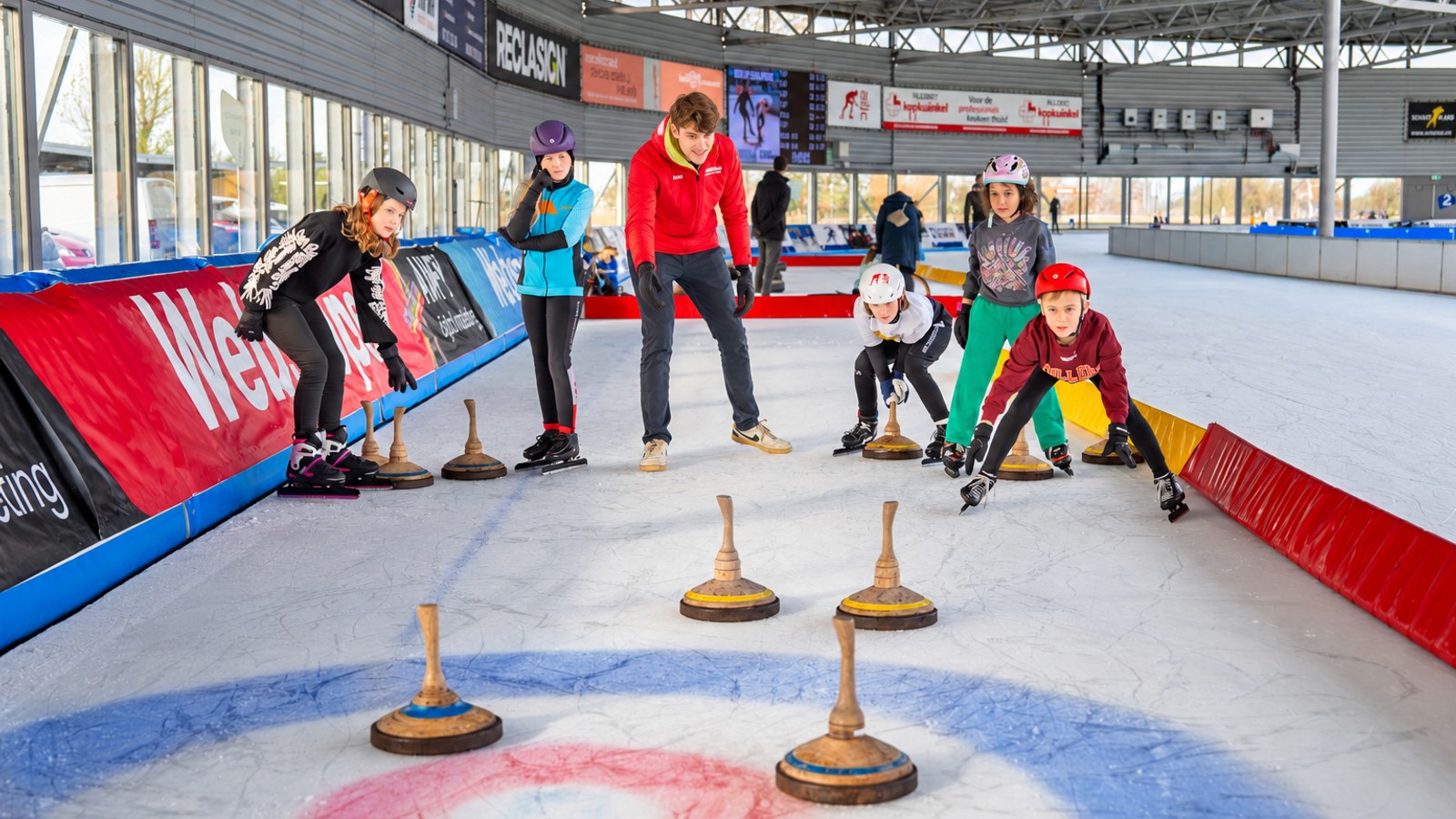 Groep kinderen en volwassenen schaatsen op indoorisjsbaan met rode en blauwe boarding en houten steunblokken