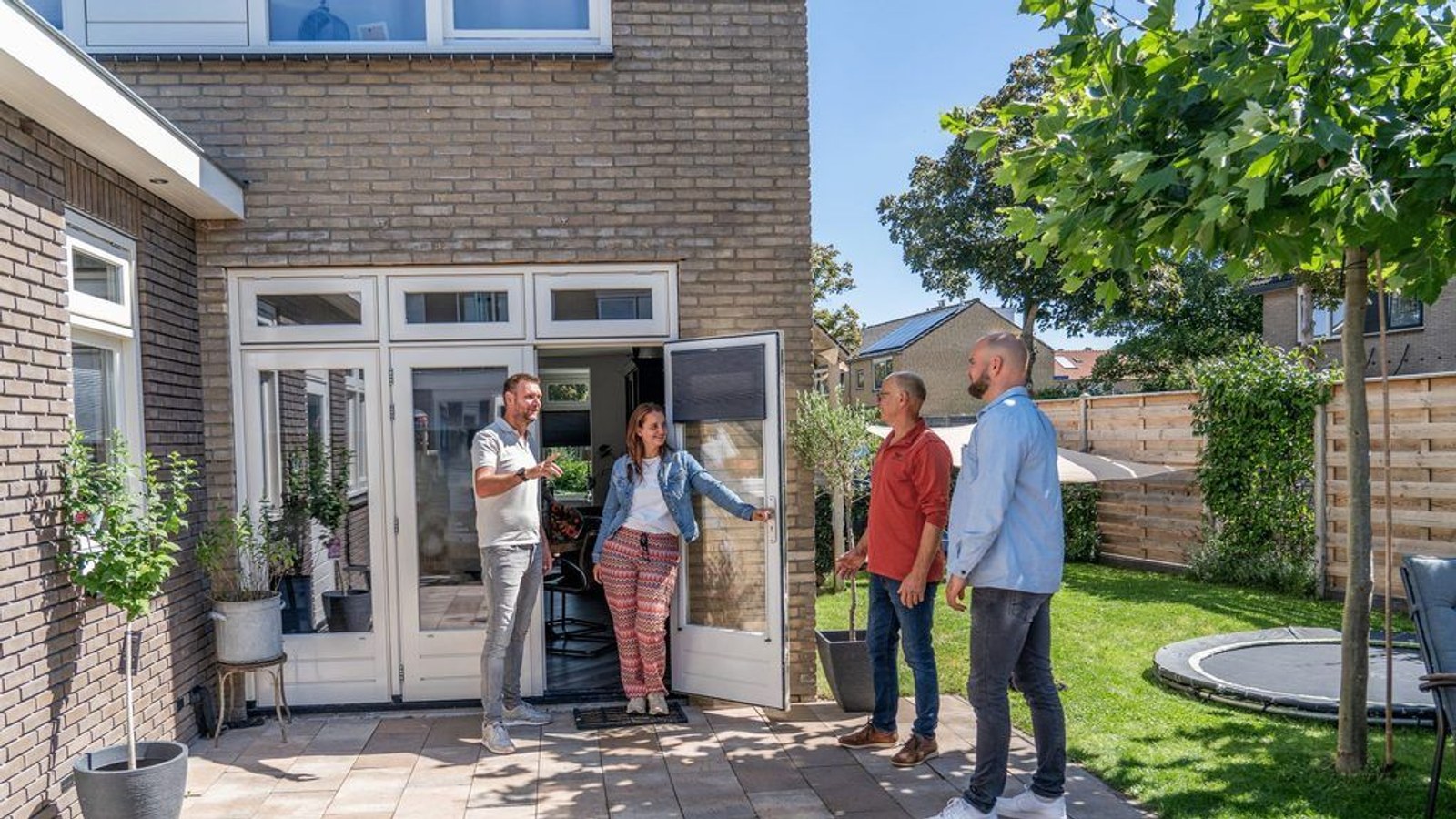 Vier personen in gesprek bij open deur van baksteenhuis met grote ramen en groene tuin