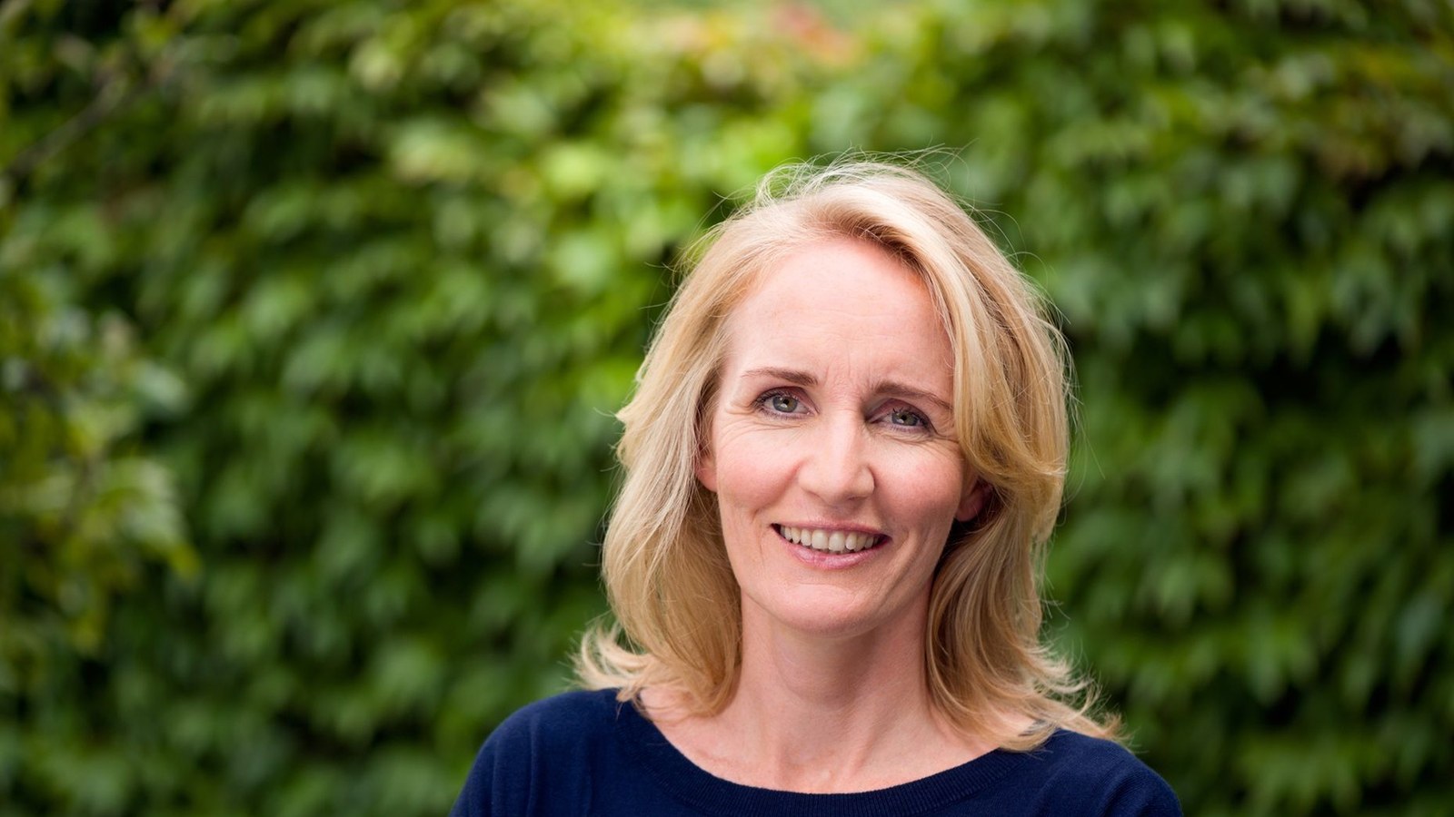 Vrouw met blond halflang haar, glimlachend naar de camera, draagt donkerblauwe trui tegen achtergrond van groen gebladerte.