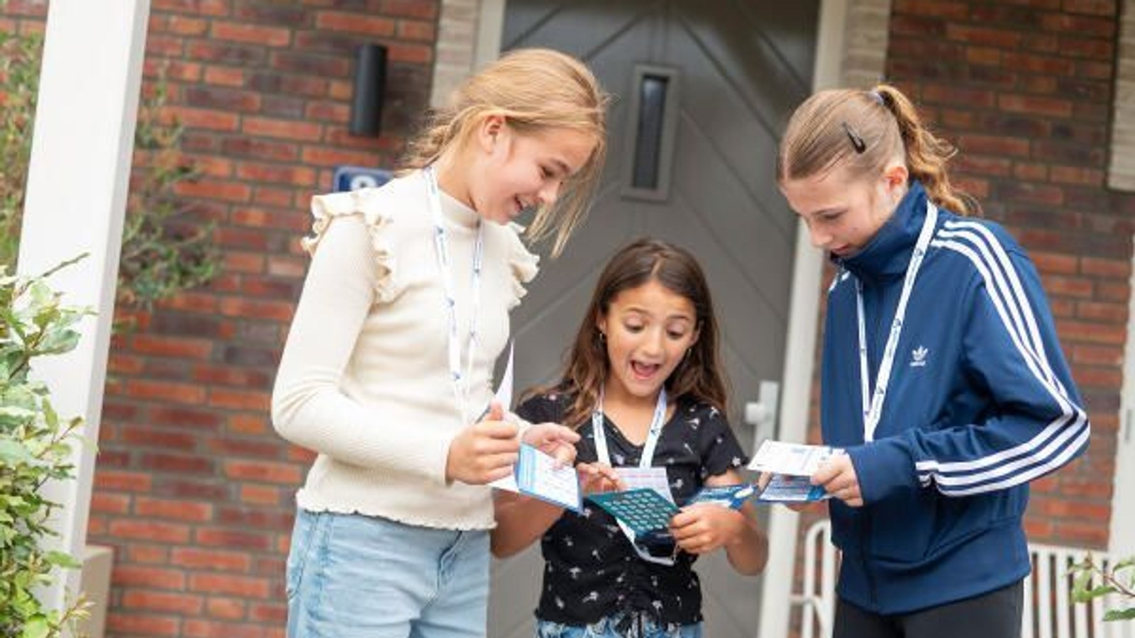 Drie meisjes bekijken samen een blauwe portemonnee of tasje voor een rode bakstenen woning met witte deur.
