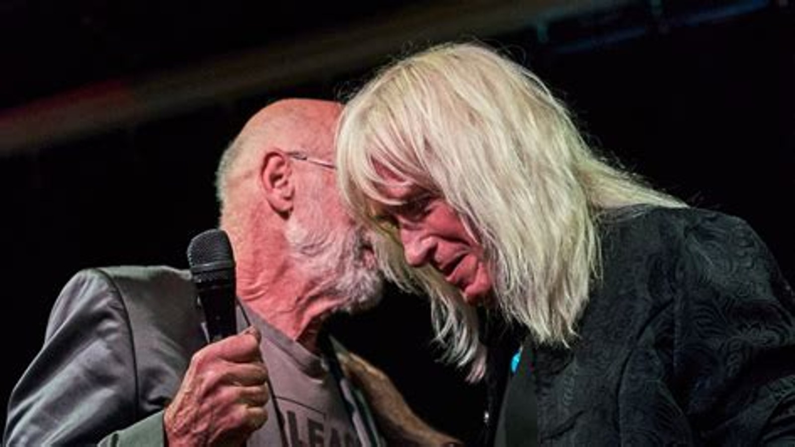 Twee mannen op podium met microfoon, een dragend wit haar en bril.