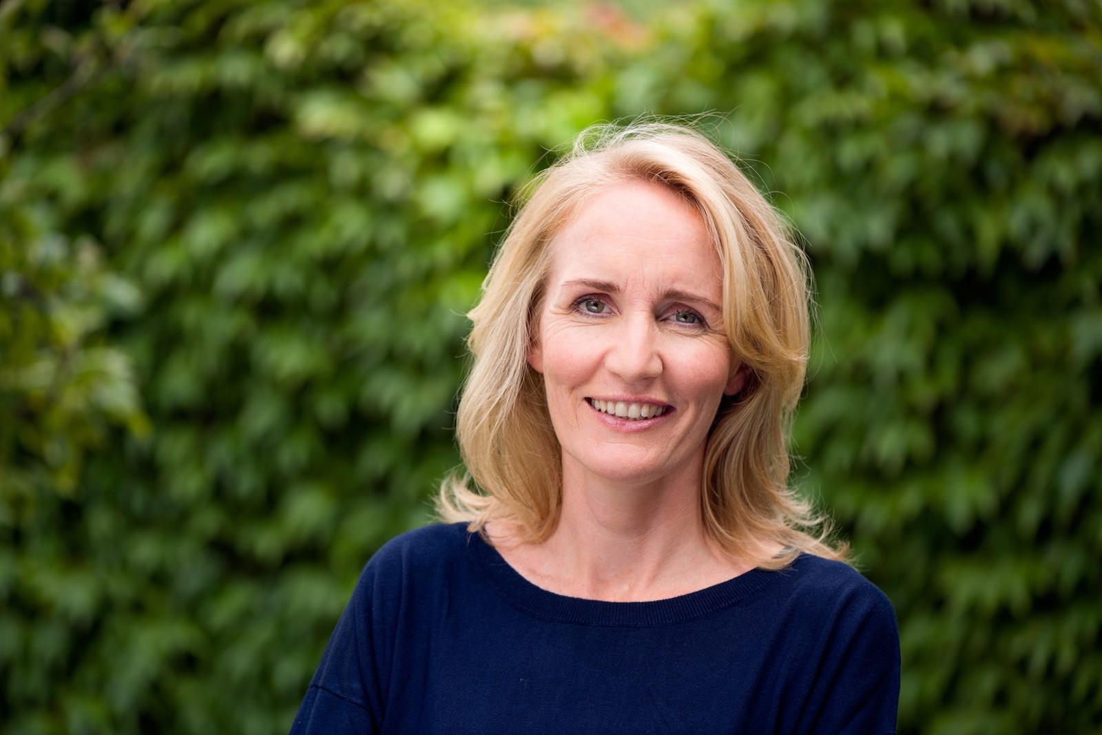 Vrouw met blond schouderlang haar, glimlachend naar camera, draagt donkerblauw shirt tegen achtergrond van groen gebladerte.