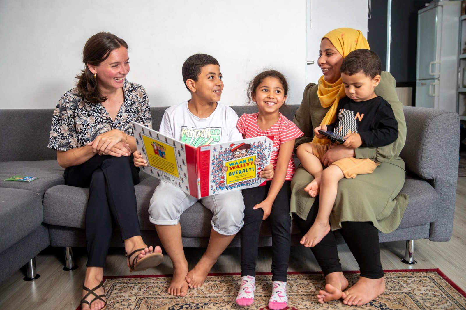 Twee volwassenen en drie kinderen zitten op een grijze bank en lezen samen uit een boek met de titel "Waar is Wally". Ze bevinden zich in ee