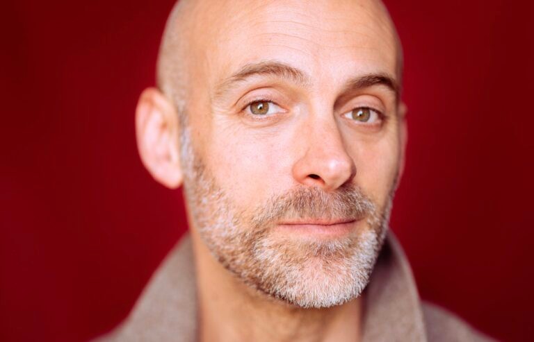 Man met kaal hoofd en grijs-bruine baard, draagt grijze trui, tegen rode achtergrond, frontaal portret.