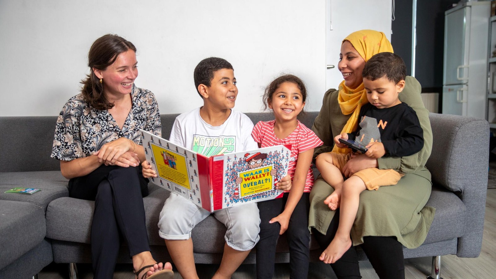 Twee vrouwen en drie kinderen zitten op een grijze bank en kijken samen naar een kinderboek met zoekplaatjes. Ze bevinden zich in een modern