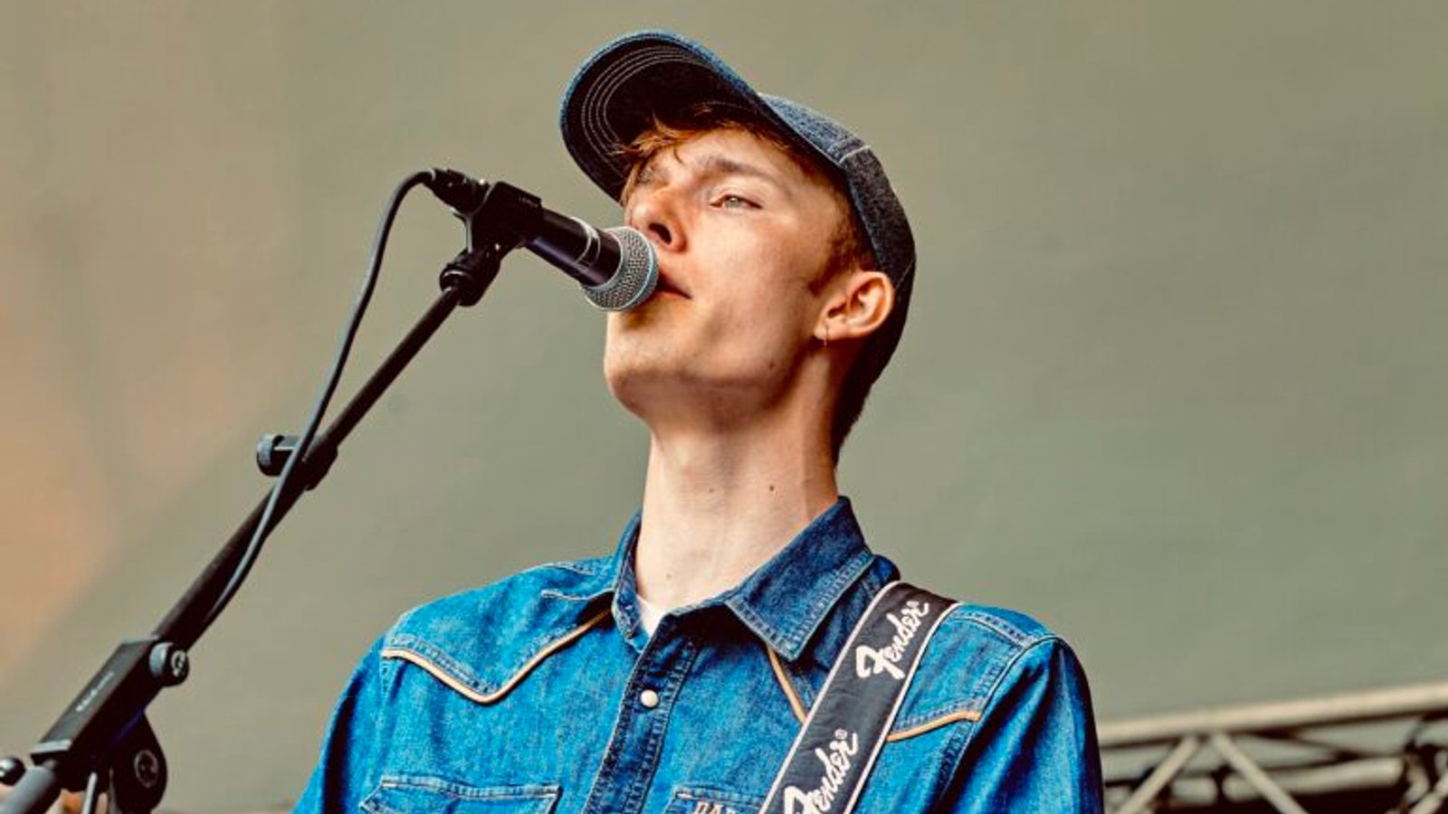 Gitarist bespeelt elektrische gitaar tijdens optreden, draagt blauw denim overhemd en zwarte gitaarsnoer.