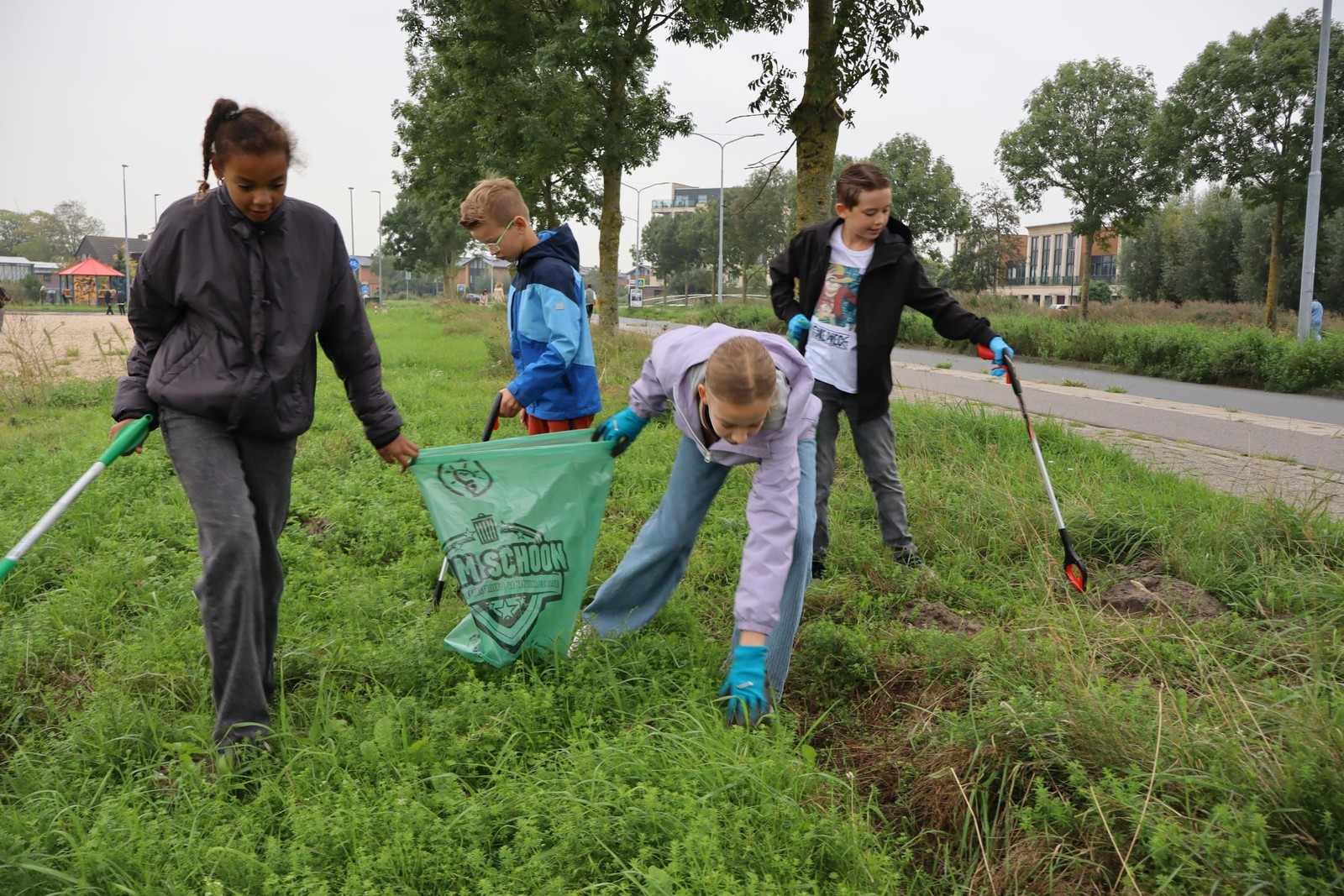 Vier kinderen en een volwassene ruimen zwerfvuil op in een groen veld langs een weg. Een kind houdt een groene zak vast met het opgeraapte a