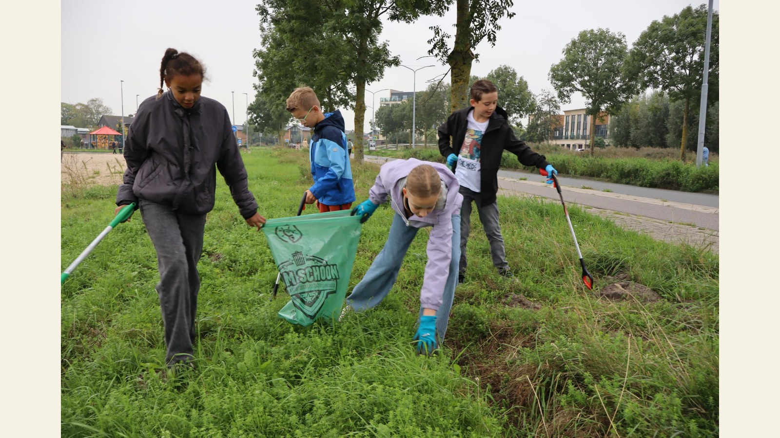 Vijf kinderen en volwassenen ruimen zwerfvuil op in een groen veld langs een weg. Ze dragen handschoenen en gebruiken opruimprikkers en groe