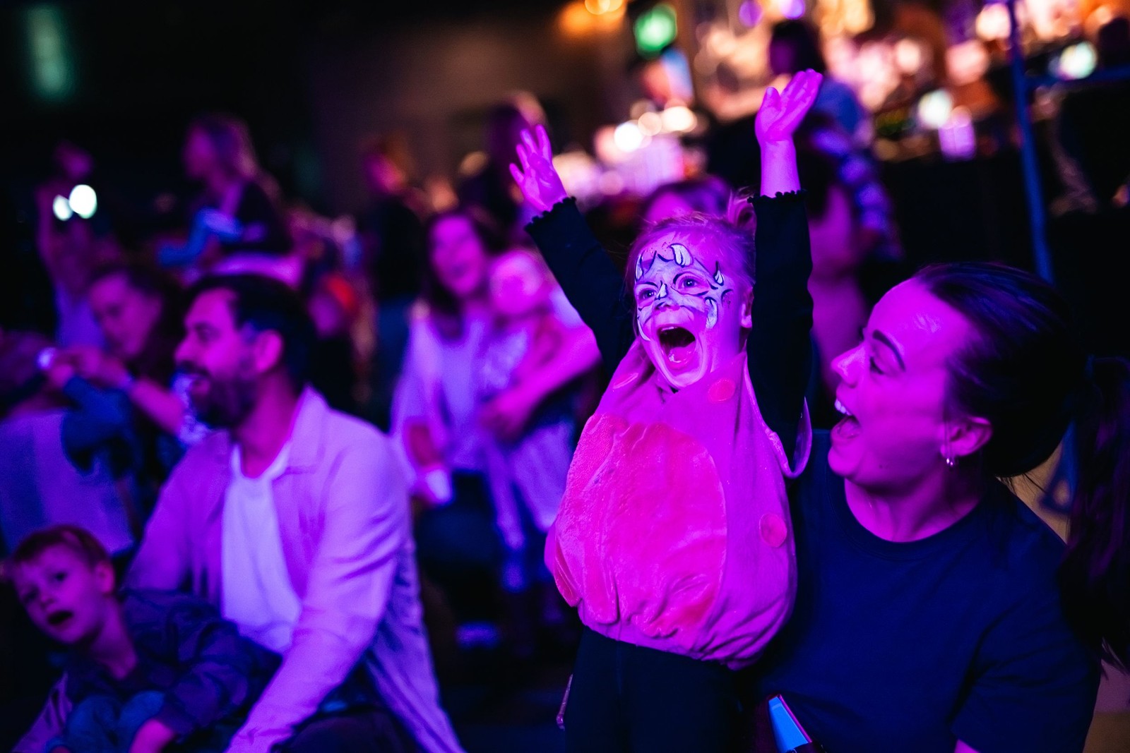 Groep mensen danst en springt op een feest met paarse en blauwe neonverlichting. Een persoon in het midden draagt gezichtsverf en een roze s
