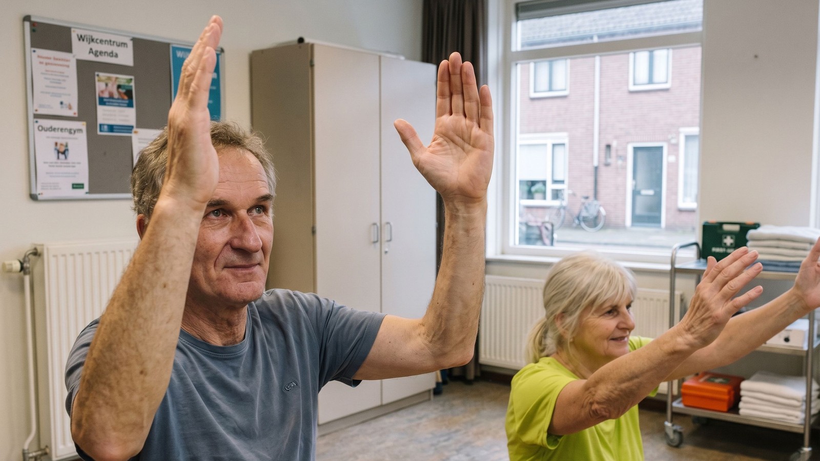 Man en vrouw voeren armbeweging uit tijdens beweegles in ontmoetingscentrum met ramen en kasten op achtergrond