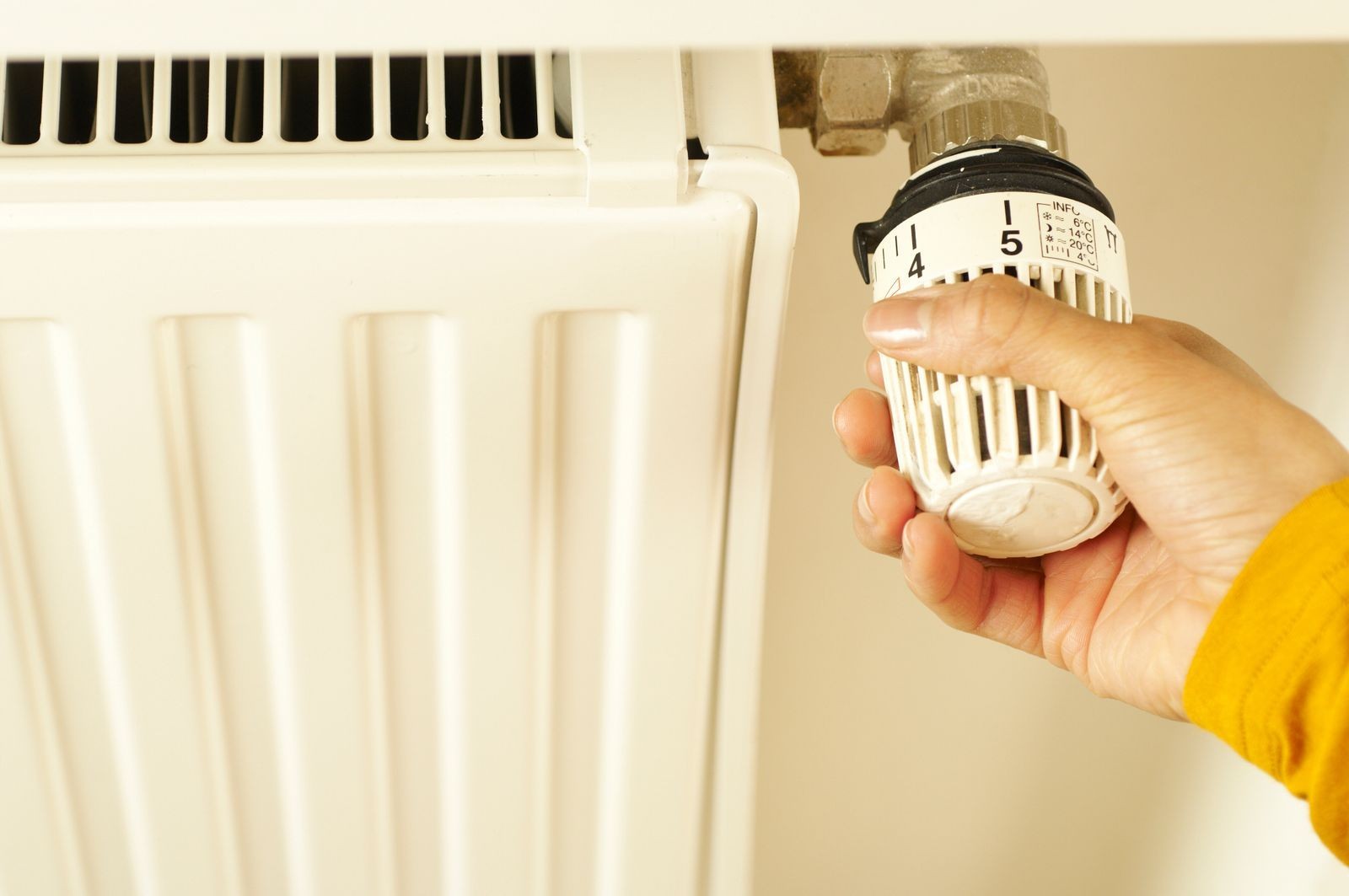 Hand stelt thermostaat van radiator in op stand 5, radiator met verticale lamellen op achtergrond, gele mouw zichtbaar.
