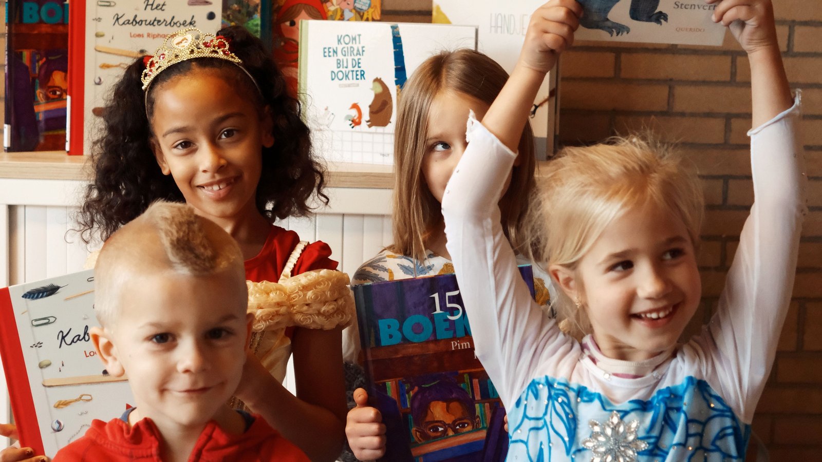Vier kinderen houden kinderboeken vast voor een bakstenen muur met een groot cartoon-karakter afgebeeld. Ze dragen feestkleding en glimlache