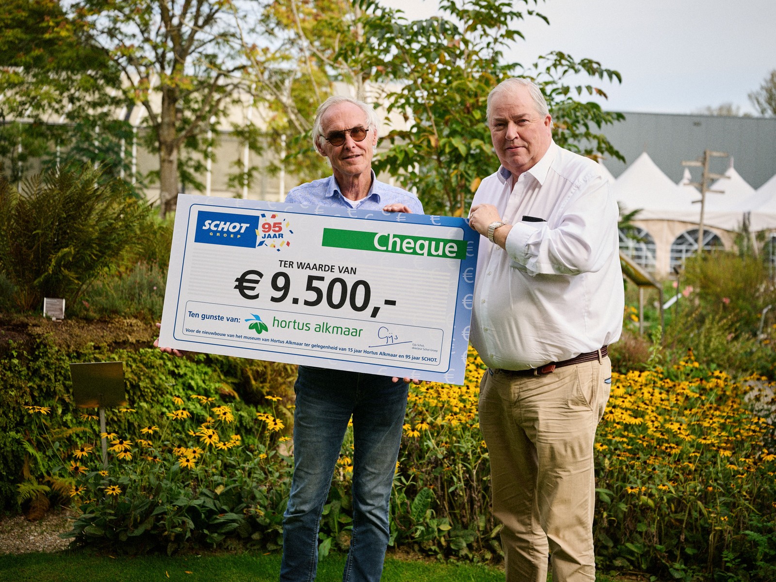 Twee mannen houden grote cheque van negen duizend vijfhonderd euro vast in kruidentuin met bloemen en struiken.