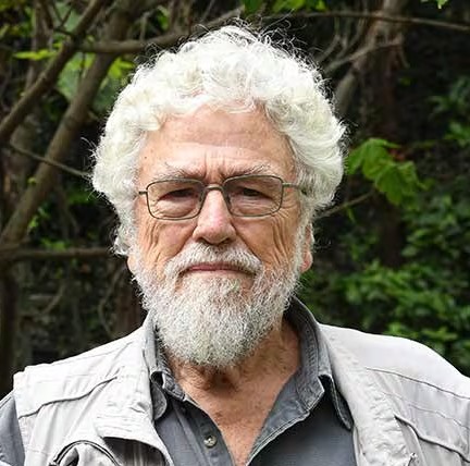 Oudere man met wit haar en baard, bril, lichtgrijze overhemd, tegen groene plantachtergrond.