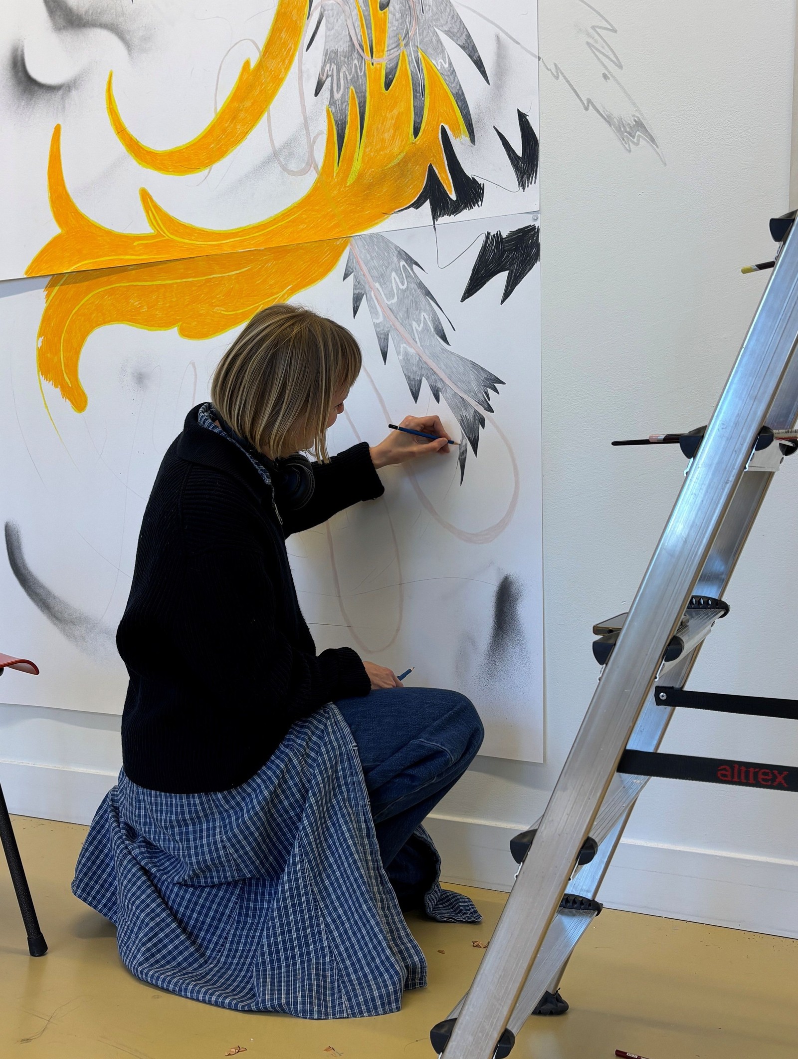 Kind knielt voor een witte wand en tekent met een viltstift aan een kunstwerk met gele en grijze vormen. Een aluminium ladder staat ernaast.