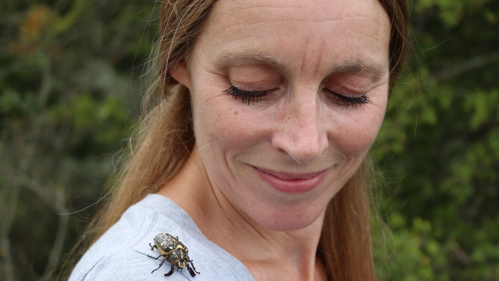 Vrouw met rood haar en gesloten ogen, glimlachend, met een insect op haar schouder tegen groene achtergrond.