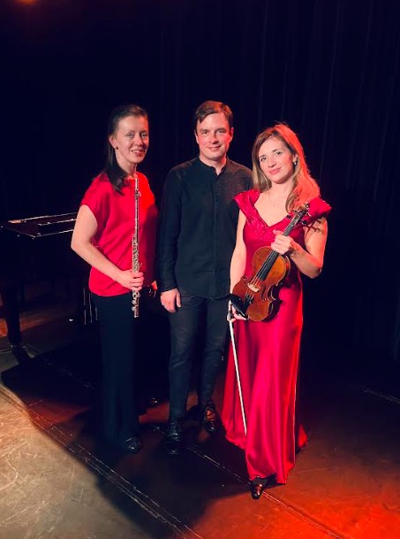 Drie musici op een podium: links een vrouw met fluit in rode blouse, midden een man in zwart shirt, rechts een vrouw met viool in rode jurk.