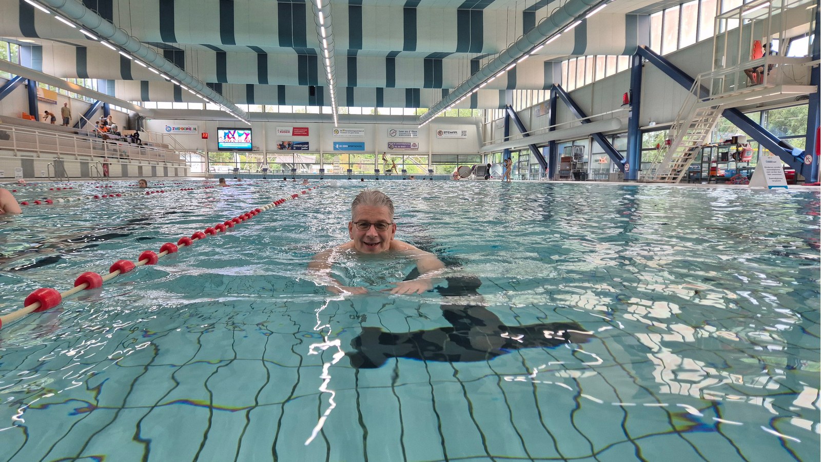 Zwemmer in een overdekt zwembad tijdens een trainingssessie, met rode drijvers die de baantjes afscheiden. Zicht op de architectuur van het
