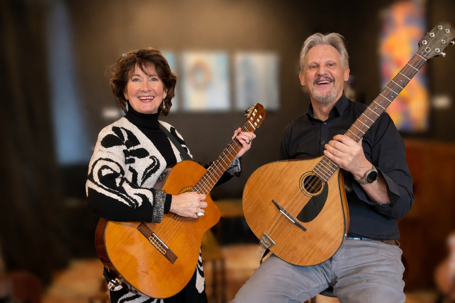 Twee personen met elk een klassieke gitaar poseren samen in een interieurruimte met onscherpe achtergrond. De persoon links draagt een zwart