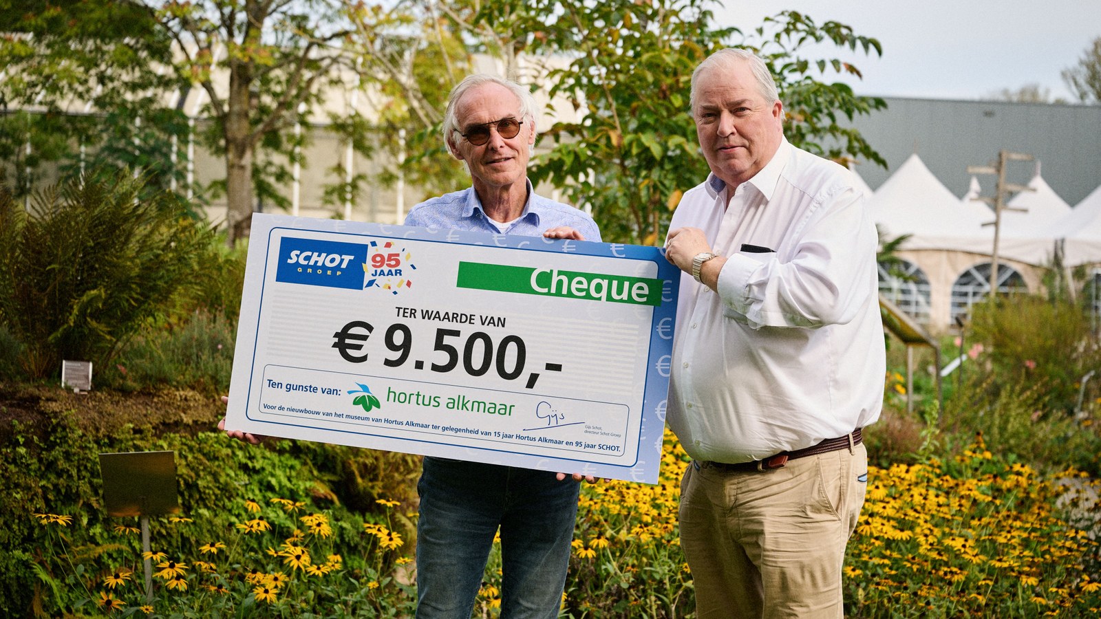 Twee mannen houden grote cheque van negen duizend vijfhonderd euro vast in kruidentuin met groene planten.