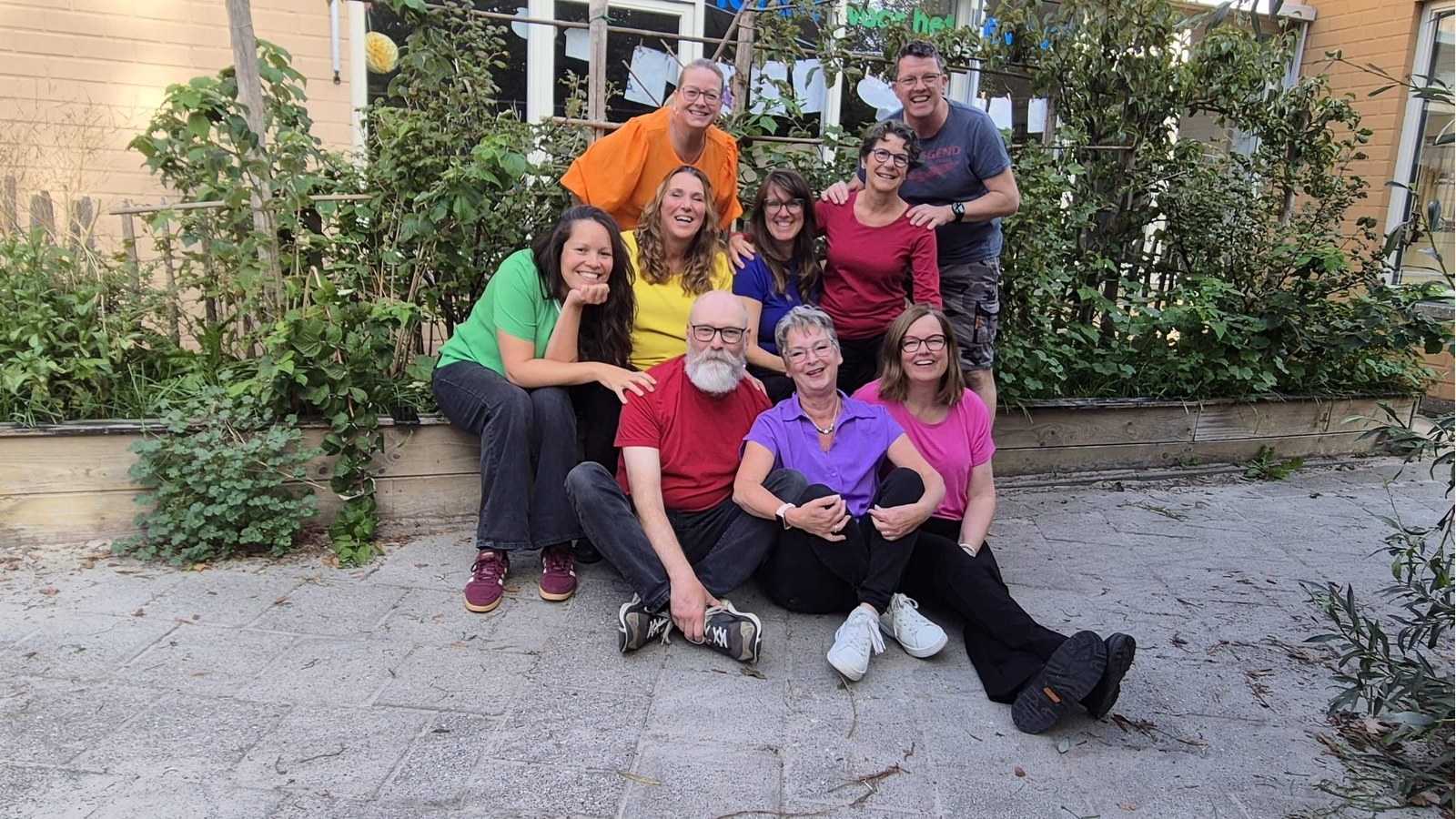 Groep van negen personen poserend in een tuin. Vier personen zitten vooraan op de grond, vijf staan erachter. Ze dragen kleding in verschill