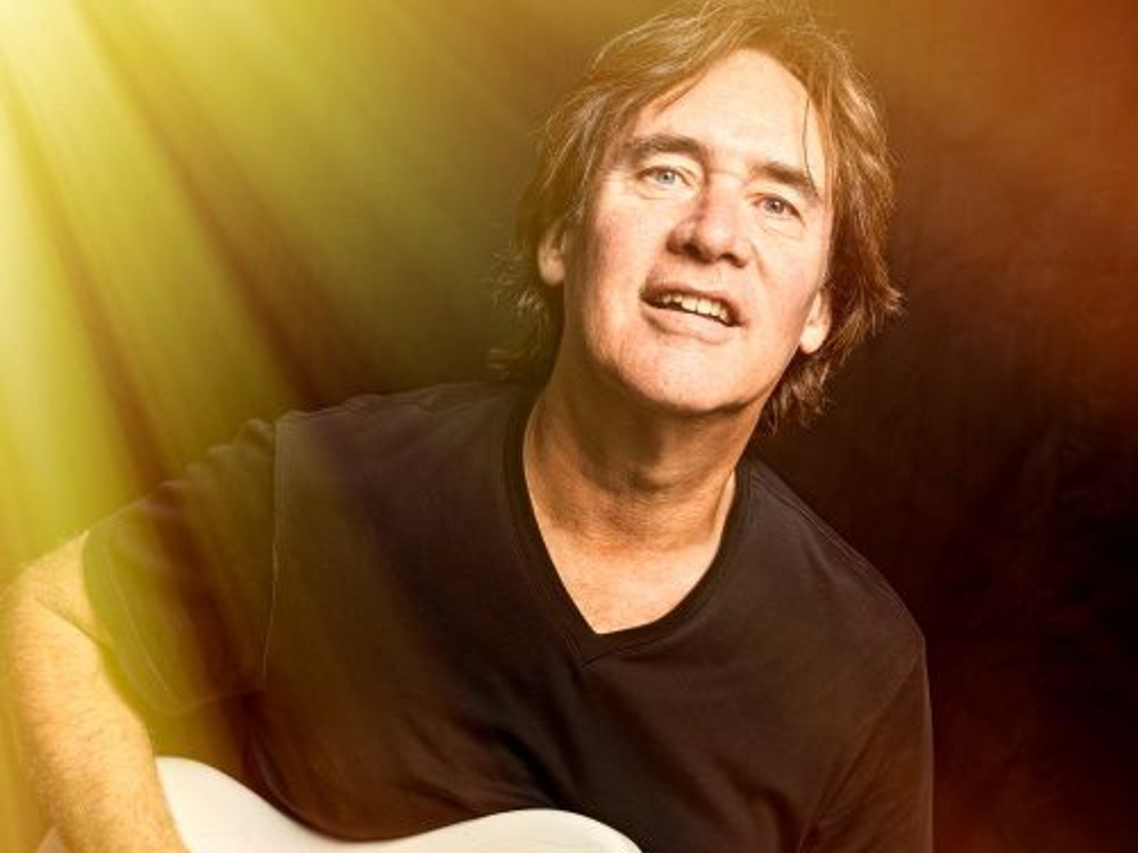 Man met halflang bruin haar en zwart shirt speelt gitaar tegen geel-oranje licht