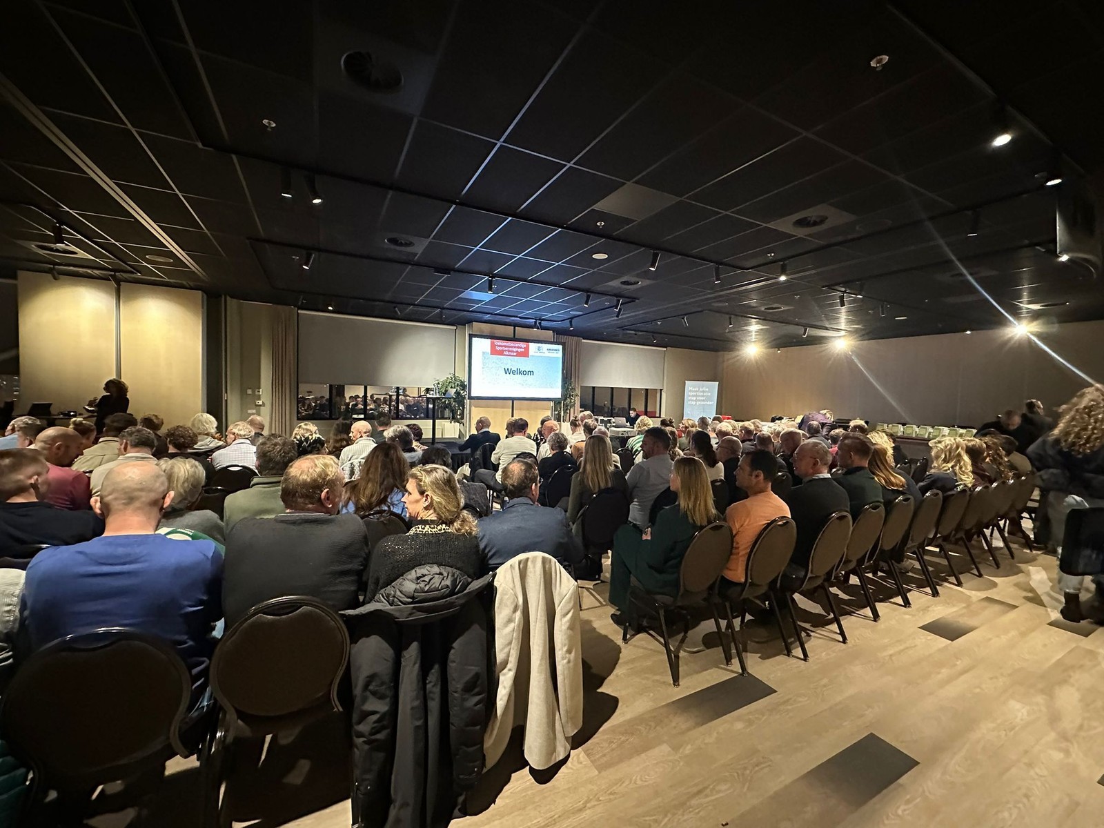 Zaal vol bezoekers zittend op stoelen tijdens een presentatie. Op het podium achter in de zaal staat een scherm met "Welkom" weergegeven. Mo