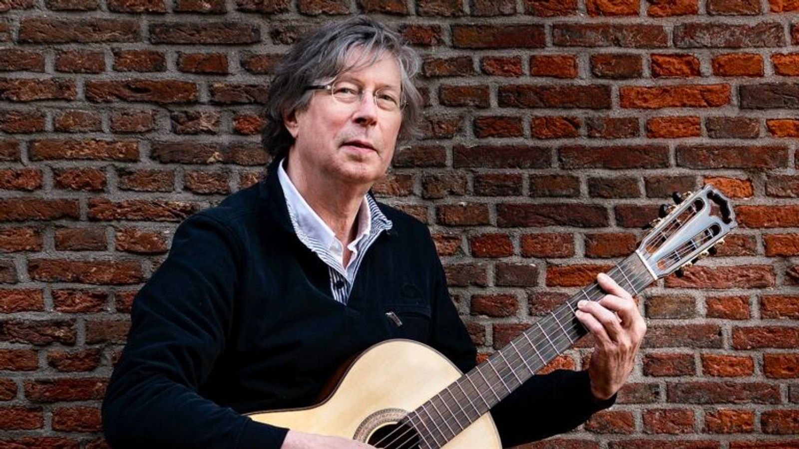 Man met grijs haar en bril speelt akoestische gitaar tegen een bakstenen muur. Hij draagt een donker vest over een gestreept overhemd.