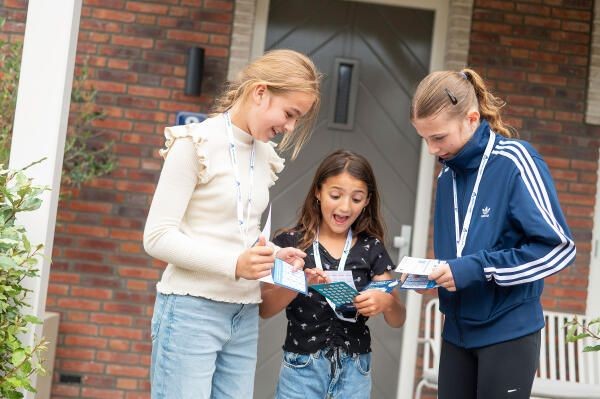 Drie meisjes bekijken samen een kaart of brochure voor een bakstenen huis. Twee dragen lanyards met badges. Een draagt een witte trui, een z