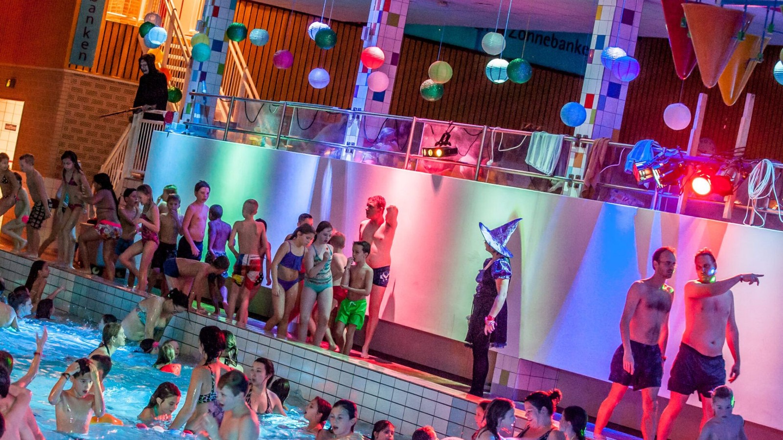 Veel kinderen zwemmen in een binnenbad met gekleurde verlichting en dansende figuren op een projectiescherm, kleurrijke ballonnen hangen