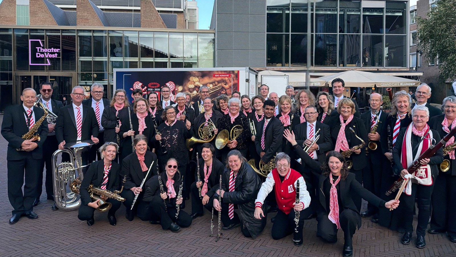 Grote groep muziekanten in zwarte pakken en roze stropdassen met blazers poseren met hun instrumenten op een plein voor moderne gebouwen.