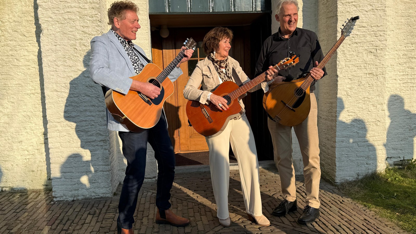 Drie muzikanten met akoestische gitaren spelen buiten voor witgekalkte kerk met donkergroene deur