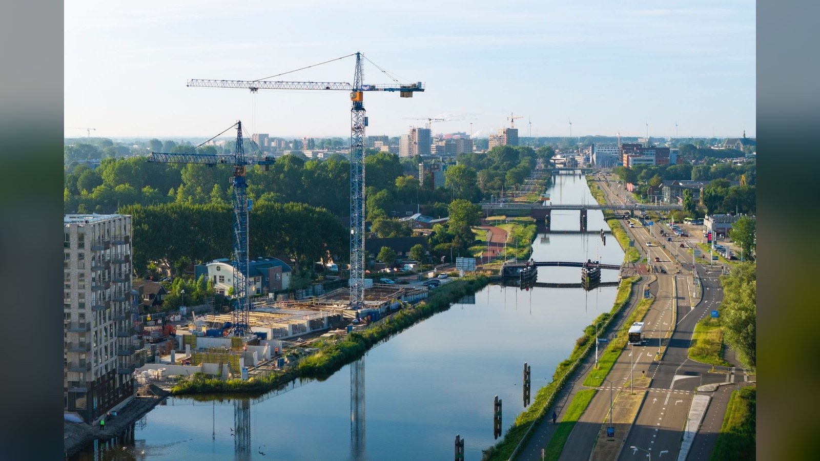 Bouwkranen boven een kanaal met een schip en werf aan de linkeroever. Rechts een weg en groene oevers. Stadsgebouwen en bomen op de achtergr