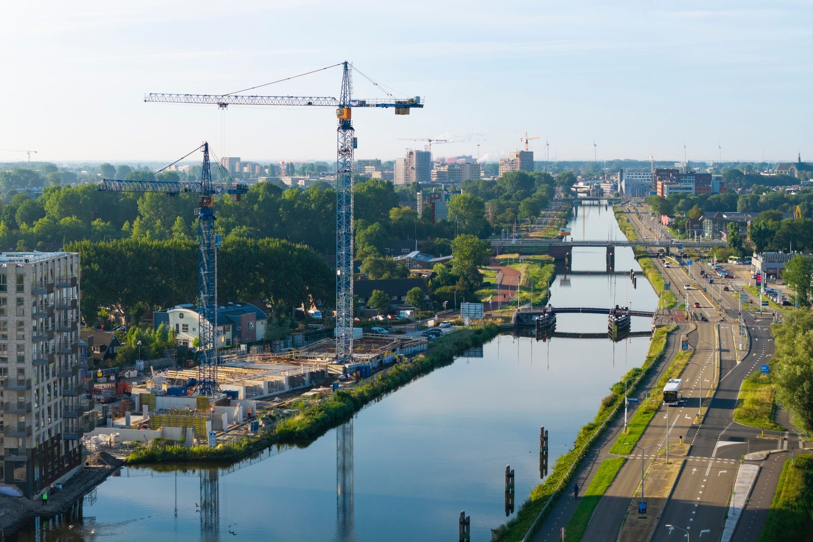 Bouwkranen en constructiewerk aan een kanaal in een stad, met een brug, groene oevers en flatgebouwen aan weerszijden van het water.