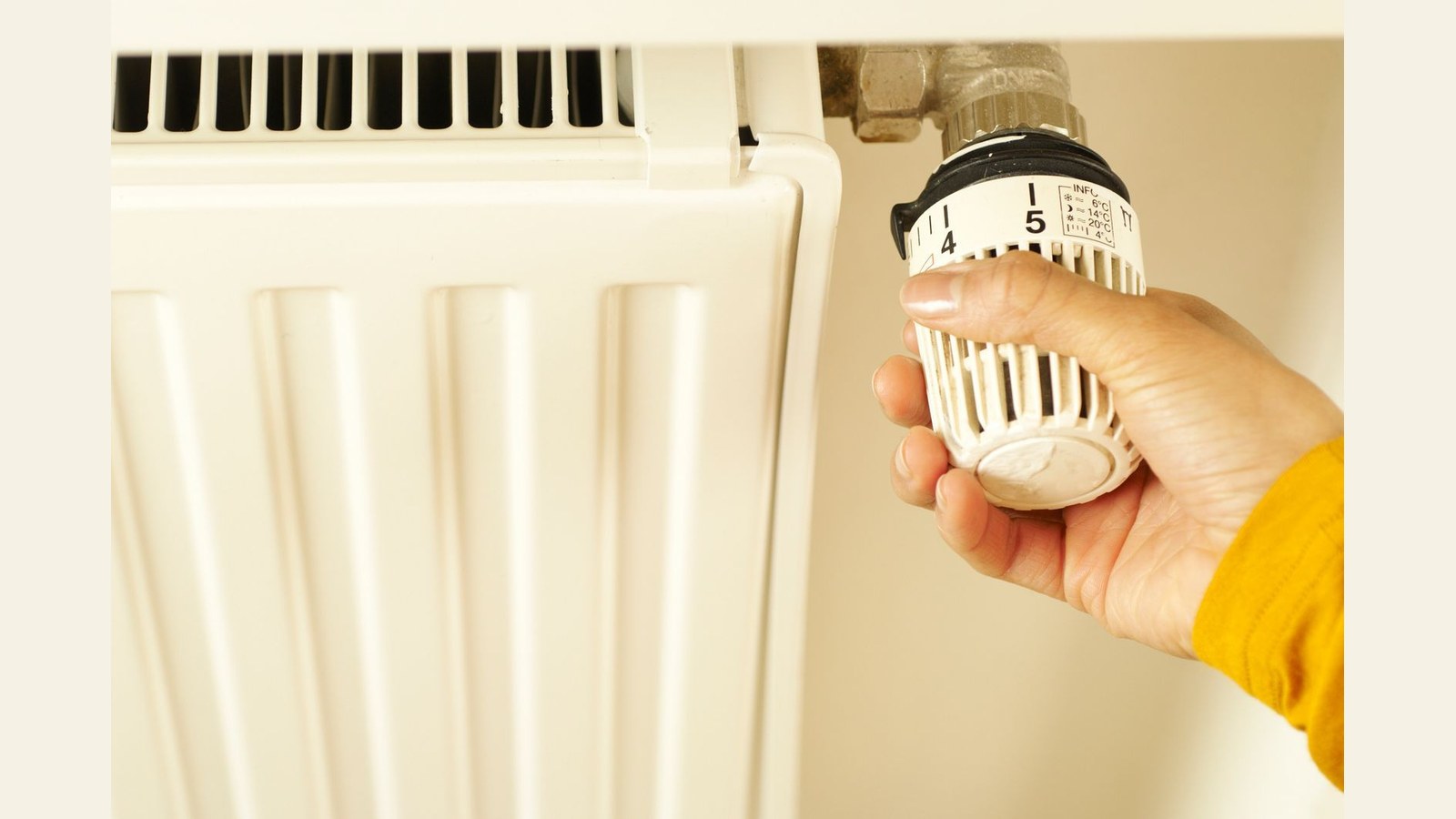 Hand in geel shirt stelt thermostaat van radiator bij op nummer 5, witte radiator zichtbaar op achtergrond.
