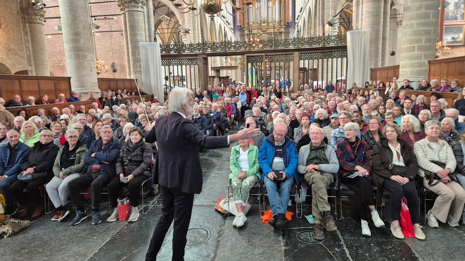 Man in zwart pak spreekt tot grote groep mensen zittend in kerkinterieur met stenen zuilen en balkon op achtergrond.